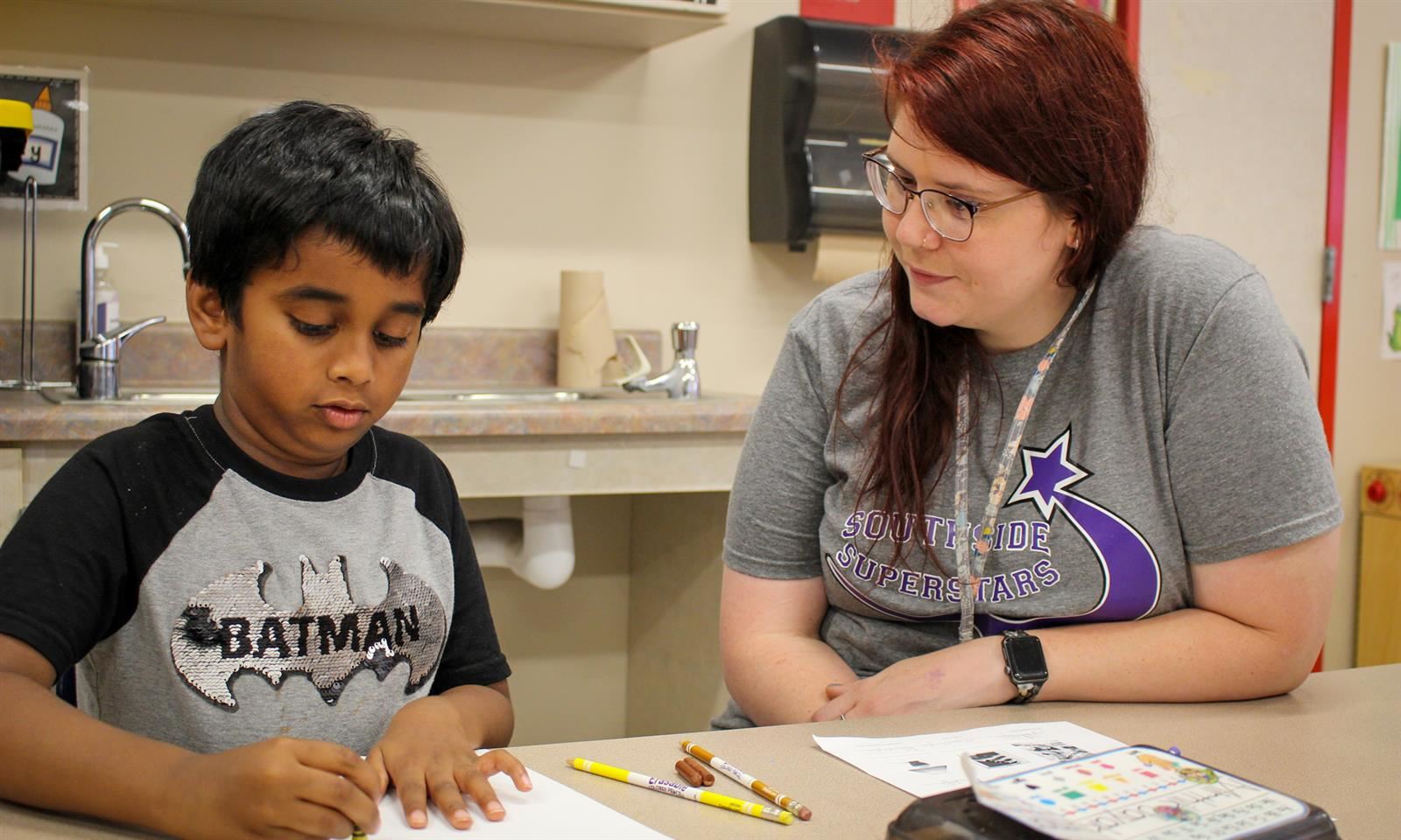 A teacher watches a student fill out a handout while they sit at a table